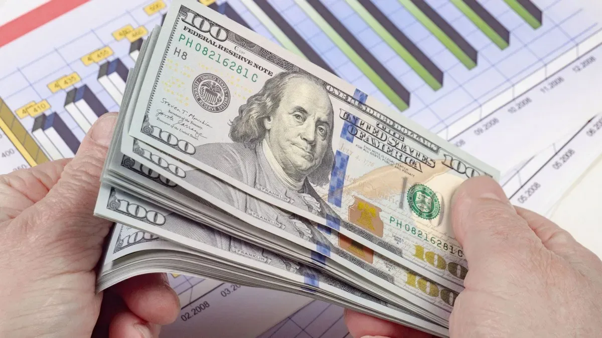 Billetes de dólares estadounidenses apilados junto a una cotización digital en pantalla