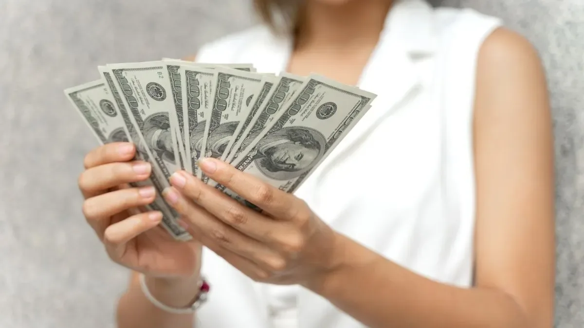 Persona sosteniendo billetes de dólar frente a una pantalla con cotizaciones cambiarias