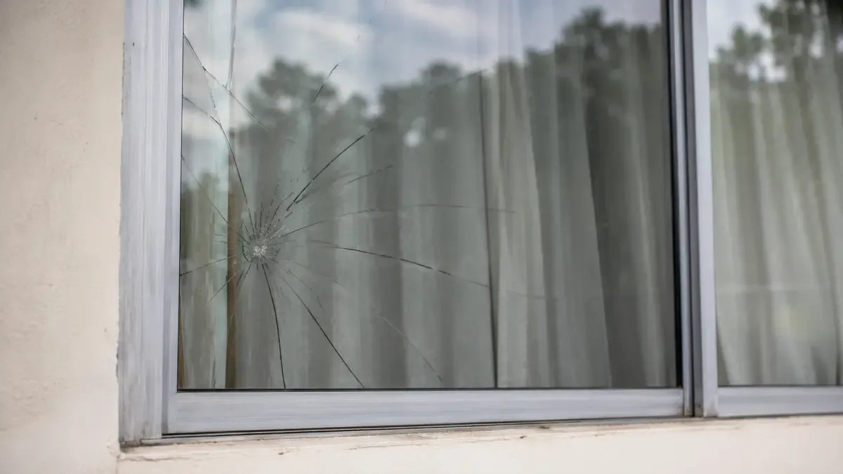 Ventana de una casa con el vidrio quebrado en forma de telarana y una cortina detras